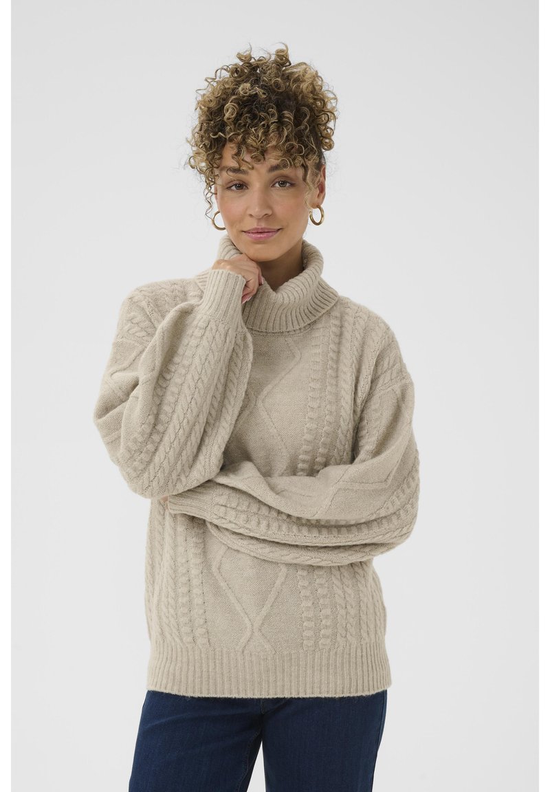 Beige kaapeknitted turtleneck-swearer med brede ærmer, strukturerte mønstre og ribbestrikket kant. Kombinert med mørke jeans på en nøytral bakgrunn.