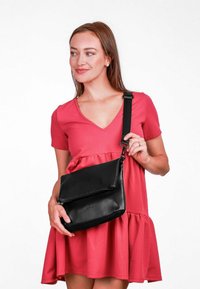 Černá kožená taška na rameno s klopou a detailem na zip, obsahující odnímatelný černý popruh. Hladká textura, jednoduchý design.