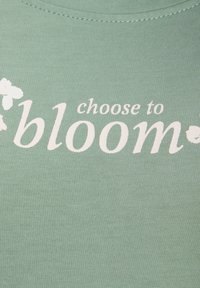 Šviesiai žalia medžiaga su baltais užrašu „choose to bloom“ ir mažais gėlių piešiniais abiejose užrašo pusėse.