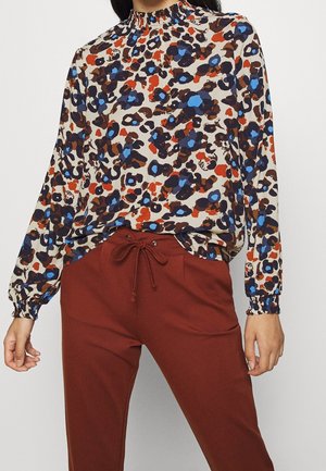 Vrouw draagt een blouse met lange mouwen en een hoge hals met een blauw, bruin en oranje bloemenprint, ingestopt in roestkleurige broek met trekkoord.