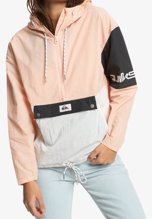 Woman wearing a pastel pink, white, and black windbreaker with a half-zip, hood, drawstrings, and light blue jeans, hand in pocket.