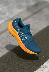 Blauwe hardloopschoen met oranje zool, voorzien van een gestructureerde bovenkant, witte details en een slank ontwerp. Licht boven een oppervlak zwevend.