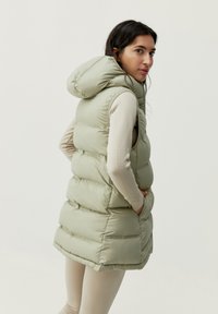 Ljusgrön vadderad väst med huva, med ett quiltat mönster och sidofickor. Tyget verkar slätt och vattentåligt.