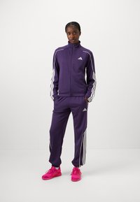 Violet treniruoklių kostiumas su baltais dryžiais ant rankovių ir šonų, pagamintas iš glotnaus audinio. Pora su ryškiai rožinėmis sportinėmis avalynėmis.