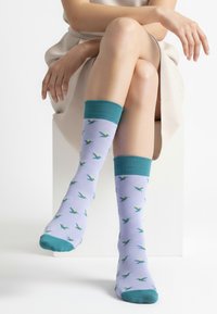 Lyse lilla sokker med en turkis kant og tå, med et mønster af små grønne og gule fugle fordelt over stoffet.