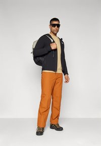 Juoda užtraukiama striukė, smėlio spalvos marškinėliai, oranžinės kelnės ir juodi žygių batai. Priedai: tamsūs akiniai nuo saulės ir pilka kuprinė.