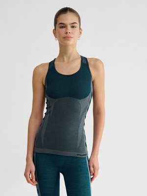 Ung kvinde iført en mørk teal tætsiddende atletisk tanktop og matchende leggings, stående mod en ensartet lys baggrund.