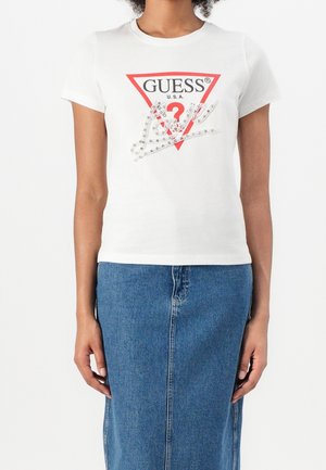 Kvinde iført hvid Guess T-shirt med korte ærmer og rød trekantlogo samt denim nederdel, stående foran en ensartet hvid baggrund.