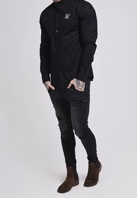 Zwart overhemd met knopen en een slim-fit, met lange mouwen en een klein geborduurd logo op de borst. Gecombineerd met versleten zwarte jeans en bruine Chelsea-laarzen.