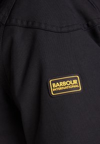 Schwarzer Stoff mit glatter Textur zeigt auf der Schulter ein rechteckiges Logo in Gelb und Schwarz mit der Aufschrift "BARBOUR INTERNATIONAL."