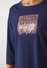 Camiseta azul-marinho com decote redondo e mangas três quartos. Apresenta uma gráfica de texto metálico em rosa dourado em um design retangular.