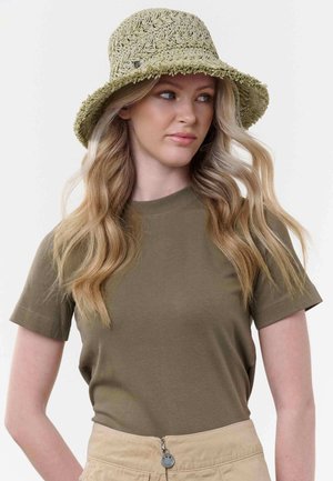 Tânără cu păr blond lung și ondulat, purtând o pălărie de tip bucket texturată bej, un tricou verde măsliniu și pantaloni bej cu talie înaltă.