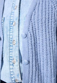 Chemise en denim bleu clair avec des boutons en argent sous un cardigan en tricot bleu clair texturé, fermé par des boutons ronds.