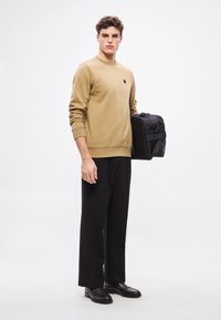 Béžová mikina s rebrovanými manžetami, nosená s čiernymi nohavicami s širokými nohavicami. Model drží čierny batoh s predným vreckom a rukoväťou.