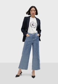 Wide-leg denim jeans in light blue with a textured surface, paired with a white graphic t-shirt and a fitted black blazer. Black heeled shoes.