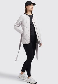 Lys grå quiltet jakke med ståkrave, frontlynlås og sidesnore. Båret over en sort top og leggings med hvide sneakers.