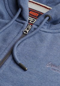 Sweat à capuche zippé bleu en tissu doux avec une finition texturée, comportant une fermeture éclair en métal, des cordons de serrage et des détails de logo brodés.