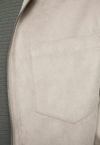 Beige faux suede-jacka med en strukturerad gråstickad insida. Har en dragkedja fram och en enda bröstficka. Mjuk, slät finish.