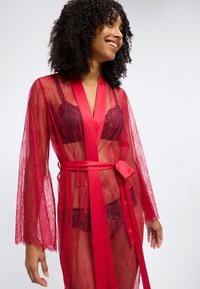 Γυναίκα που χαμογελάει φορώντας διάφανη κόκκινη δαντελένια ρόμπα δεμένη στη μέση πάνω από ταιριαστό εσώρουχο, με σγουρά μαλλιά και χαλαρή στάση.