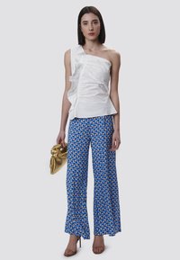 Witte asymmetrische top met ruffles, gecombineerd met blauwe wijde broek met een patroon van geometrische vormen, en houdt een gouden tas met textuur vast.
