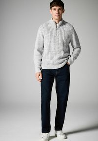 Suéter de punto trenzado gris con cuello alto, combinado con jeans oscuros y zapatillas blancas, de pie frente a un fondo neutro.