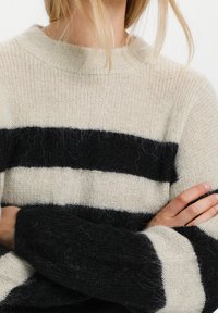 Neulottu villapaita, jossa on kerma- ja mustia vaakasuoria raitoja, ribbireunainen ympäri kaula-aukko ja teksturoitu kangas. Mukava ilme näkyvillä kuidulla.