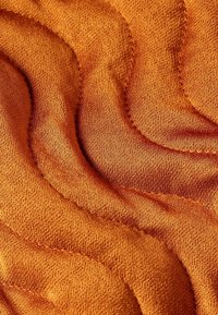 Zbliżenie na teksturowany pomarańczowy materiał z wypukłymi, falistymi wzorami przeszytymi, tworzącymi efekt pikowania.