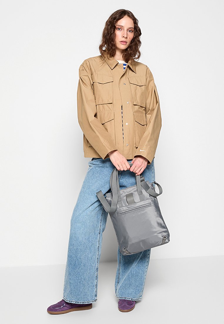 Beige leichte Jacke mit mehreren Taschen, kombiniert mit blauen Jeans im weiten Schnitt. Graue Tasche mit verstellbaren Trägern, schlichtes Design.