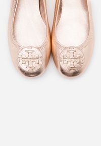 Paire de ballerines métalliques en or rose avec des emblèmes ronds du logo sur les bouts, présentée sur fond blanc.