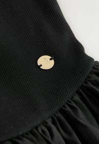 Étiquette ronde en métal doré avec la lettre « B », attachée à un tissu côtelé noir relié à un tissu noir froncé.