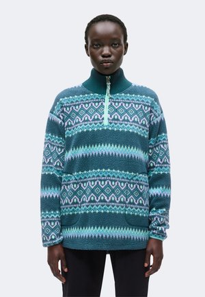 Personne portant un pull polaire à motifs bleu sarcelle avec une demi-fermeture éclair et un col montant, debout devant un fond uni gris clair.