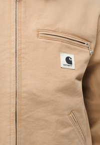 Μπεζ μπουφάν με φερμουάρ, με οριζόντια τσέπη στο στήθος, ταμπελάκι με το λογότυπο Carhartt κάτω από την τσέπη και κατωφερμένη πλαϊνή τσέπη σε λοξή θέση.