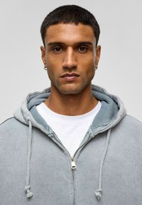 Lichtgrijze zip-up hoodie met een contrasterende lichtblauwe binnenkant, trekkoorden en een wit T-shirt eronder. De stof lijkt zacht en gestructureerd.