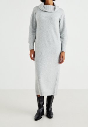 Žena v dlouhých světle šedých pletených šatech se širokým rolákem a černých kožených kozačkách po kolena, stojící na bílém pozadí.