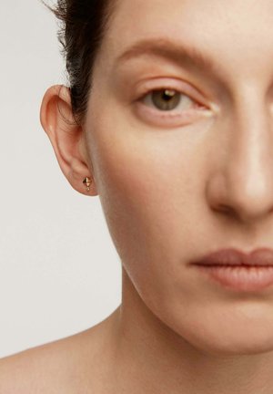 Primer plano del rostro de una persona que muestra un arete en forma de pirámide dorada con un pequeño diamante colgante en la oreja izquierda, con un fondo neutro.