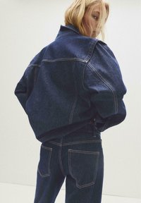 Blonde vrouw draait zich om terwijl ze een oversized donkerblauwe denimjas en bijpassende jeans draagt, tegen een effen lichte achtergrond.