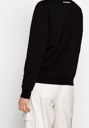 Sweatshirt noir avec des poignets et un ourlet côtelés. Logo blanc imprimé dans le dos. Le porteur l'associe avec un pantalon cargo de couleur claire.