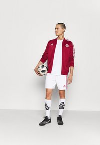 Rote Sportjacke mit weißen Akzenten, weißes T-Shirt, weiße Shorts und schwarze Fußballschuhe. Hält einen schwarz-weißen gemusterten Fuß-ball.
