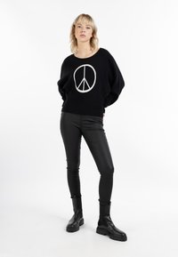 Svart tröja med en vit fredssymbol-grafik, i kombination med figursydda svarta lädershorts och svarta ankelboots, på en neutral bakgrund.
