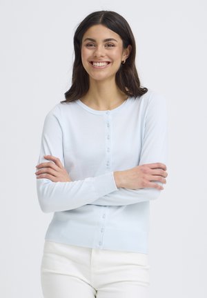 Lachende vrouw met lang donker haar, gekleed in een lichtblauwe cardigan met knopen en een witte broek, armen gekruist tegen een effen achtergrond.