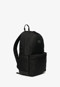 Zaino nero con spallacci imbottiti, tasca frontale con cerniera e logo "Superdry Outdoor Originals" sul comparto principale.