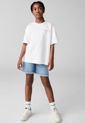 Valge ülerahuldav T-särk roosa "ALLSAINTS" logoga, kombineeritud siniste teksashortsidega, mille servad on kulunud, ja valgete spordijalatsitega, millel on mustrilised sokid.