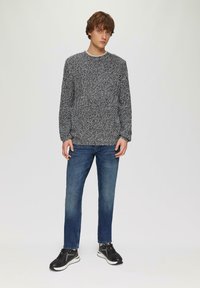 Grauer gestrickter Pullover mit strukturiertem Muster, kombiniert mit blauen Jeans und schwarzen Sportschuhen. Das Modell steht mit entspannten Armen an den Seiten.