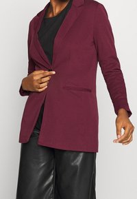 Maron blazer i texturerat tyg, enkelknapplåsning, skurna slag och sidfickor, i kombination med svarta läderbyxor.