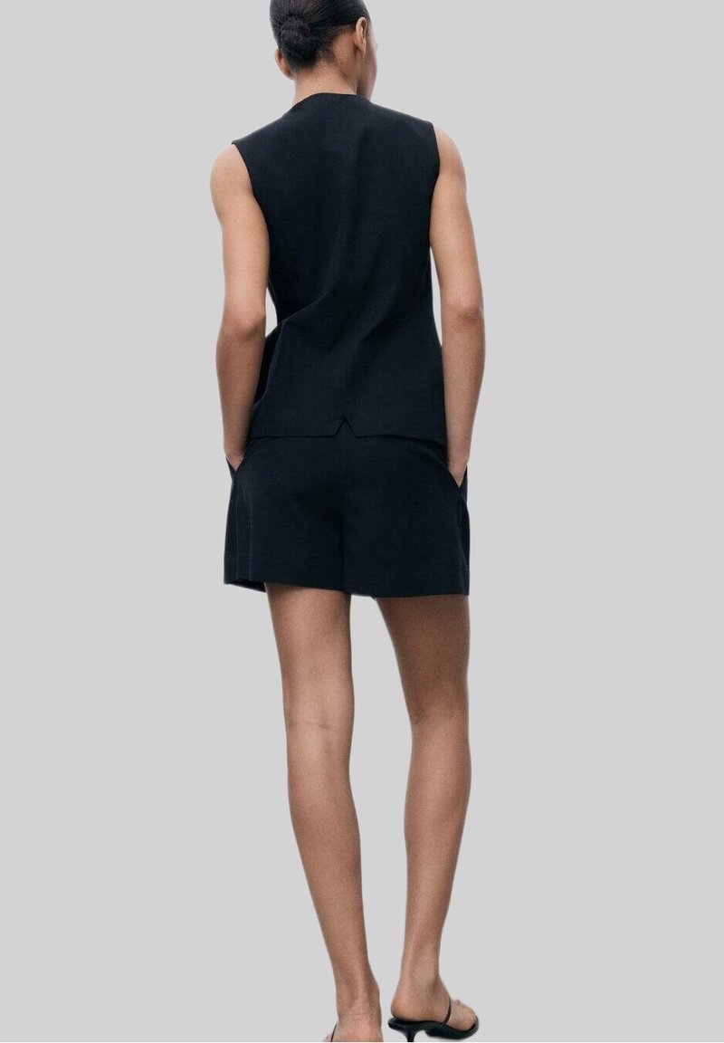 Personne avec les cheveux en chignon portant un haut noir sans manches et un short noir, debout les mains dans les poches sur un fond uni.