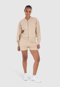 Sweatshirt en polaire beige avec fermeture éclair et shorts assortis, présentant des détails de ourlet côtelés, porté avec des baskets blanches. Design simple et décontracté.