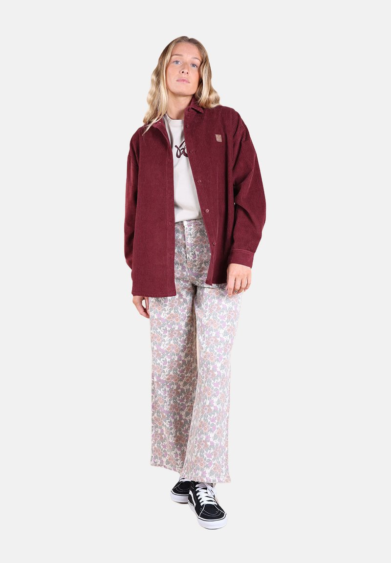 Chemise en velours côtelé bordeaux, t-shirt graphique gris, pantalon large floral dans des couleurs pastel, baskets noires avec des lacets blancs, se tenant devant un fond uni.