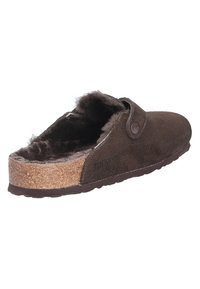 Bruine suède pantoffel met een kurken zool, voorzien van een fluffy binnenvoering en een zijsluiting met een knop voor eenvoudig aan- en uittrekken.