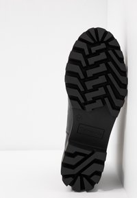Semelle de botte noire avec un motif de crampons profond et angulaire, présentée en position verticale contre un coin de mur blanc.