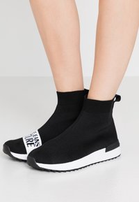Black sock-style sneakers with a knitted upper, white sole, and a decorative strap featuring "JEANS FUTURE" text. Features a pull tab at the back.